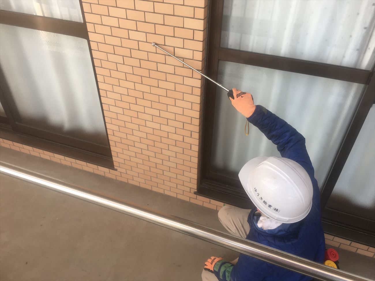 ヘルメットをかぶった作業員が、打診棒で建物の外壁タイルを点検している様子